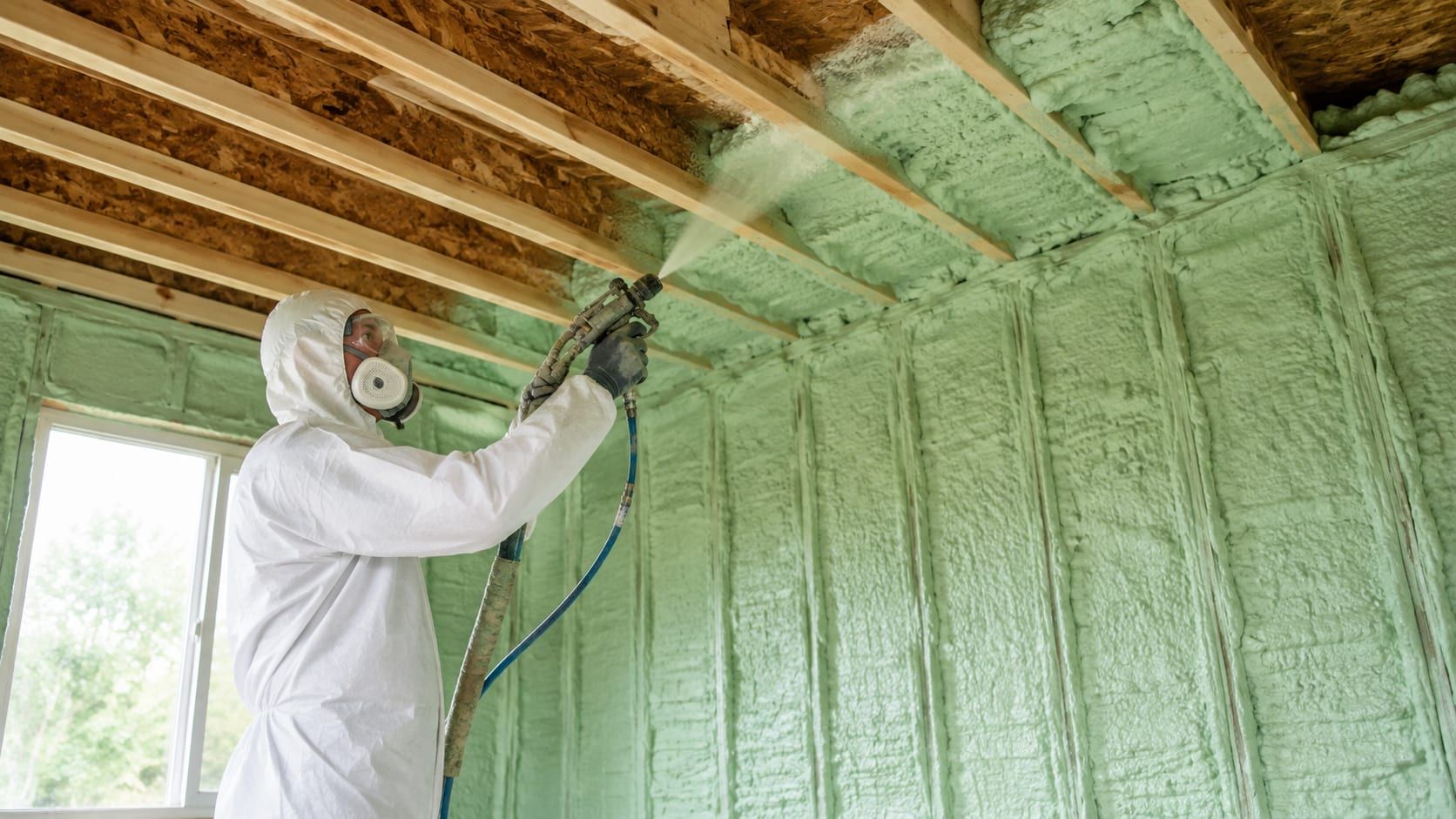 Insulation installer in a white protective suit and respirator spraying expanding foam between the wall studs and ceiling joists of a partly built room.
